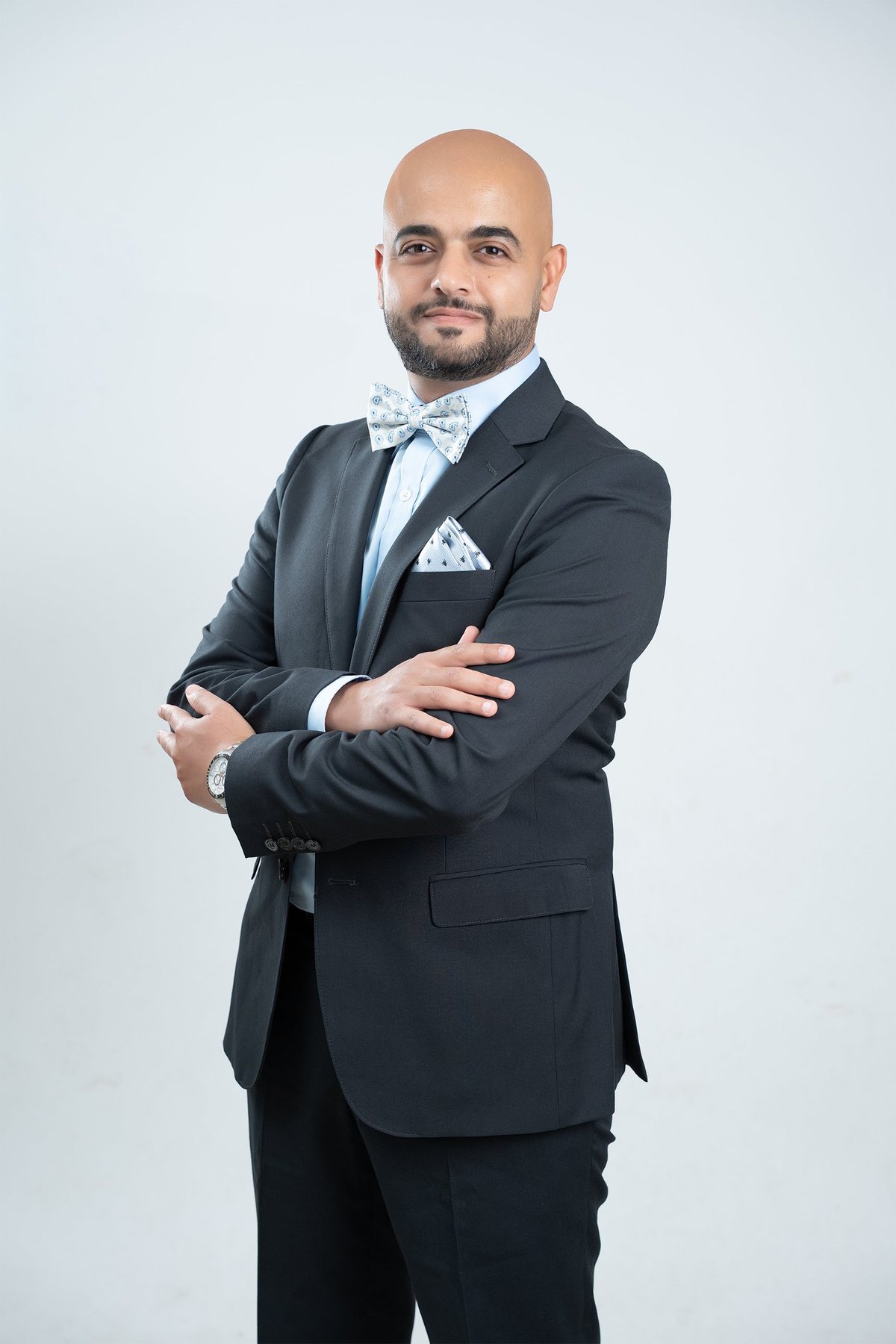 Corporate headshot — man in a charcoal suit, pale blue shirt and patterned bow tie, arms crossed and smiling on a light grey studio backdrop — executive headshots Melbourne