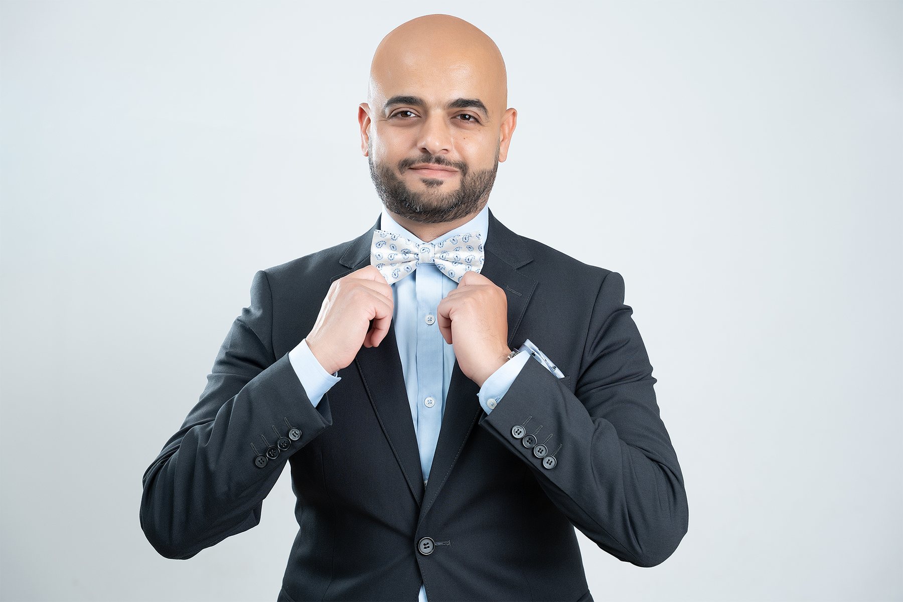 Personal branding portrait — man adjusting a white paisley bow tie, charcoal suit, soft grey backdrop — business headshots Melbourne