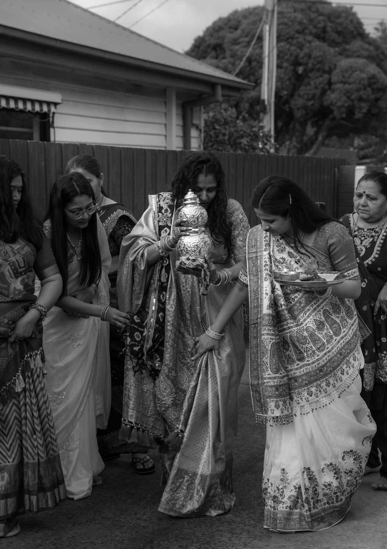 Black and white — mother-to-be carries a silver kalash, surrounded by family, at a Godh Bharai ceremony in Melbourne