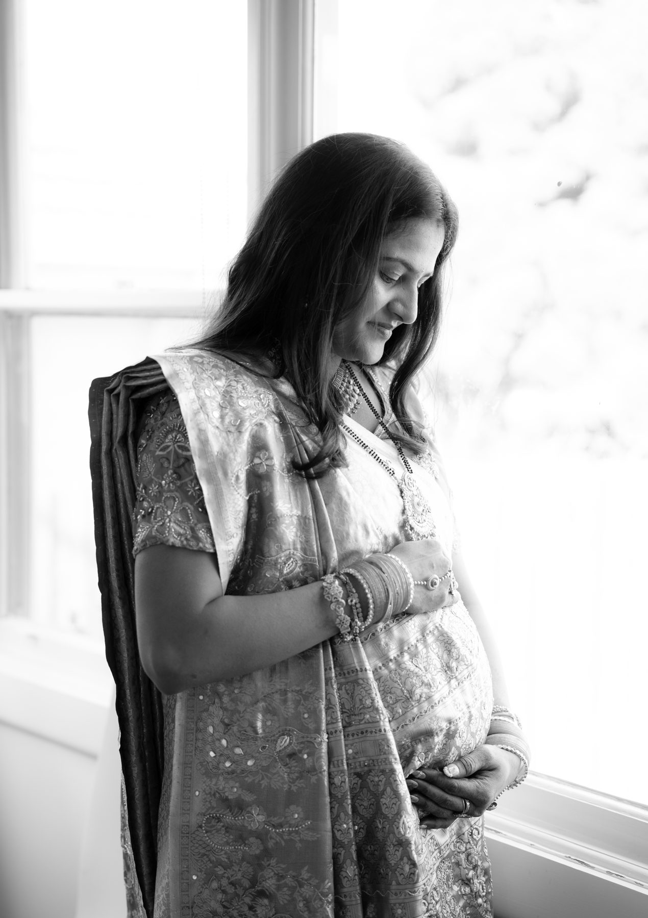 Black and white — mother-to-be in an embroidered sari cradles her bump by a window — maternity portrait Melbourne