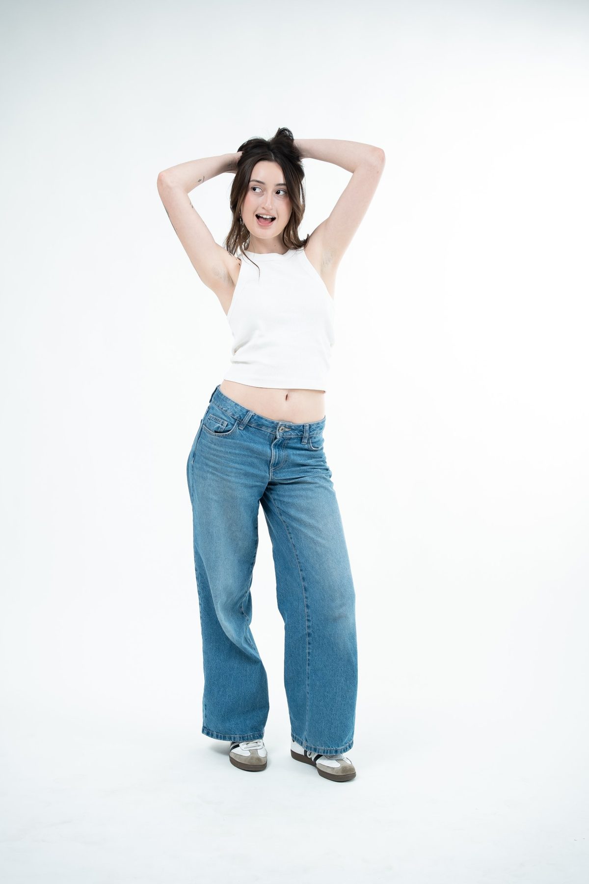 Model in a white ribbed tank and wide-leg denim, hands in her hair, glancing away with a playful expression — studio fashion lookbook, Melbourne