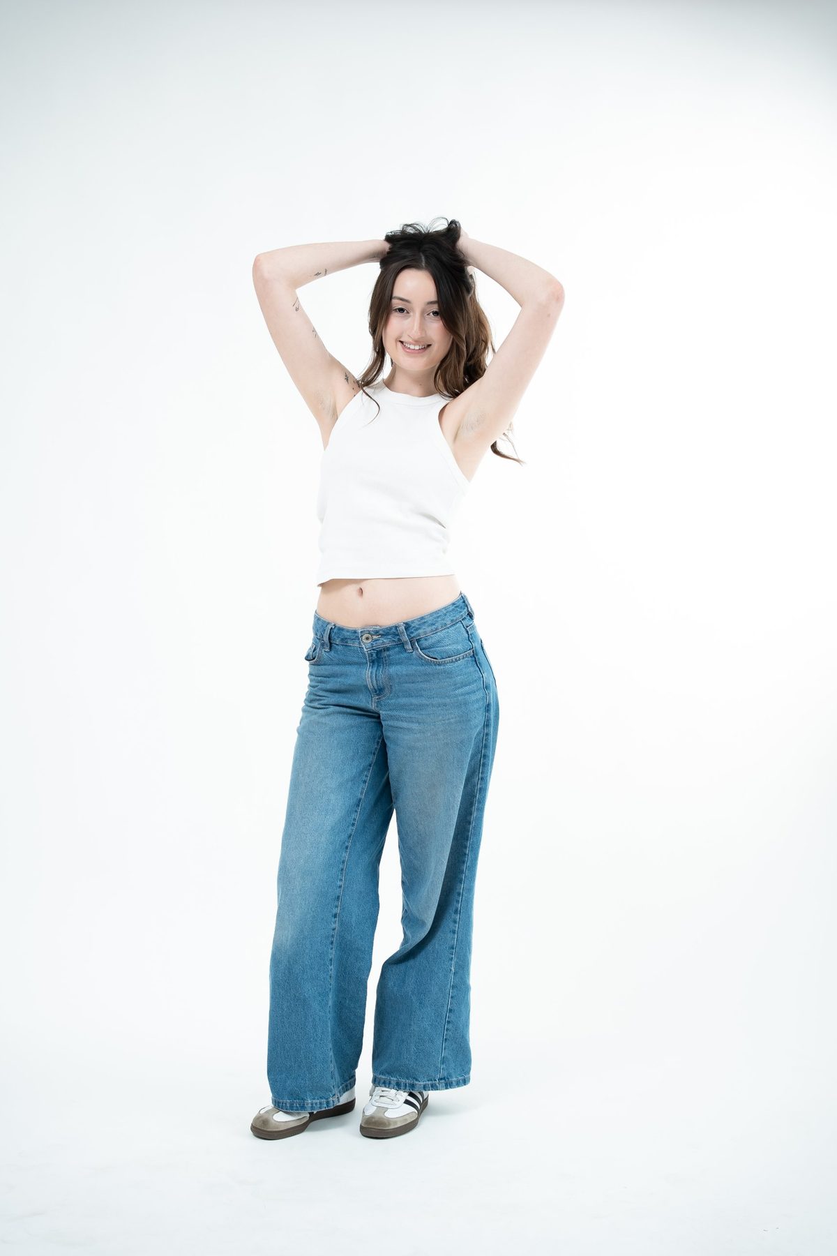 Full-length studio portrait in a white tank and wide-leg jeans, hands in her hair and smiling — white seamless backdrop, Fraser Rise studio