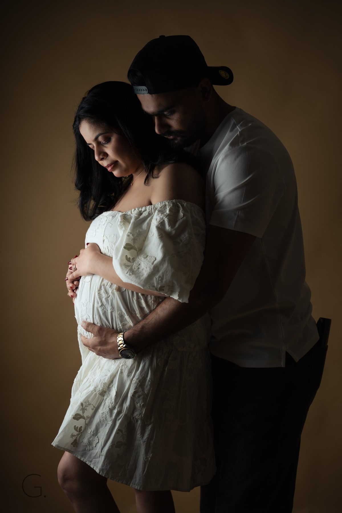 Expecting couple in a dark, low-key embrace, hands resting on the bump, white lace dress — maternity photography Melbourne