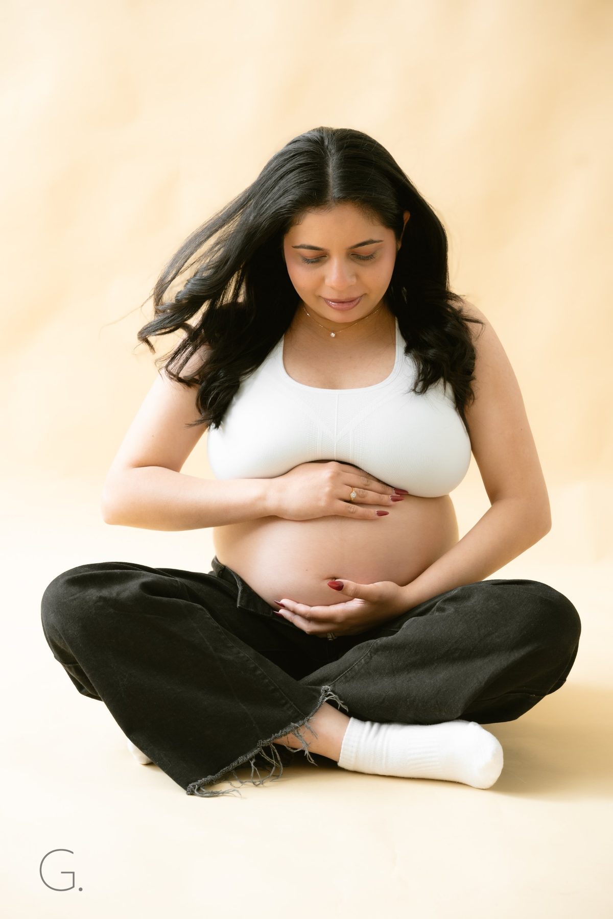Mother-to-be seated cross-legged on a warm cream backdrop, cradling her bump — studio maternity portrait, Fraser Rise