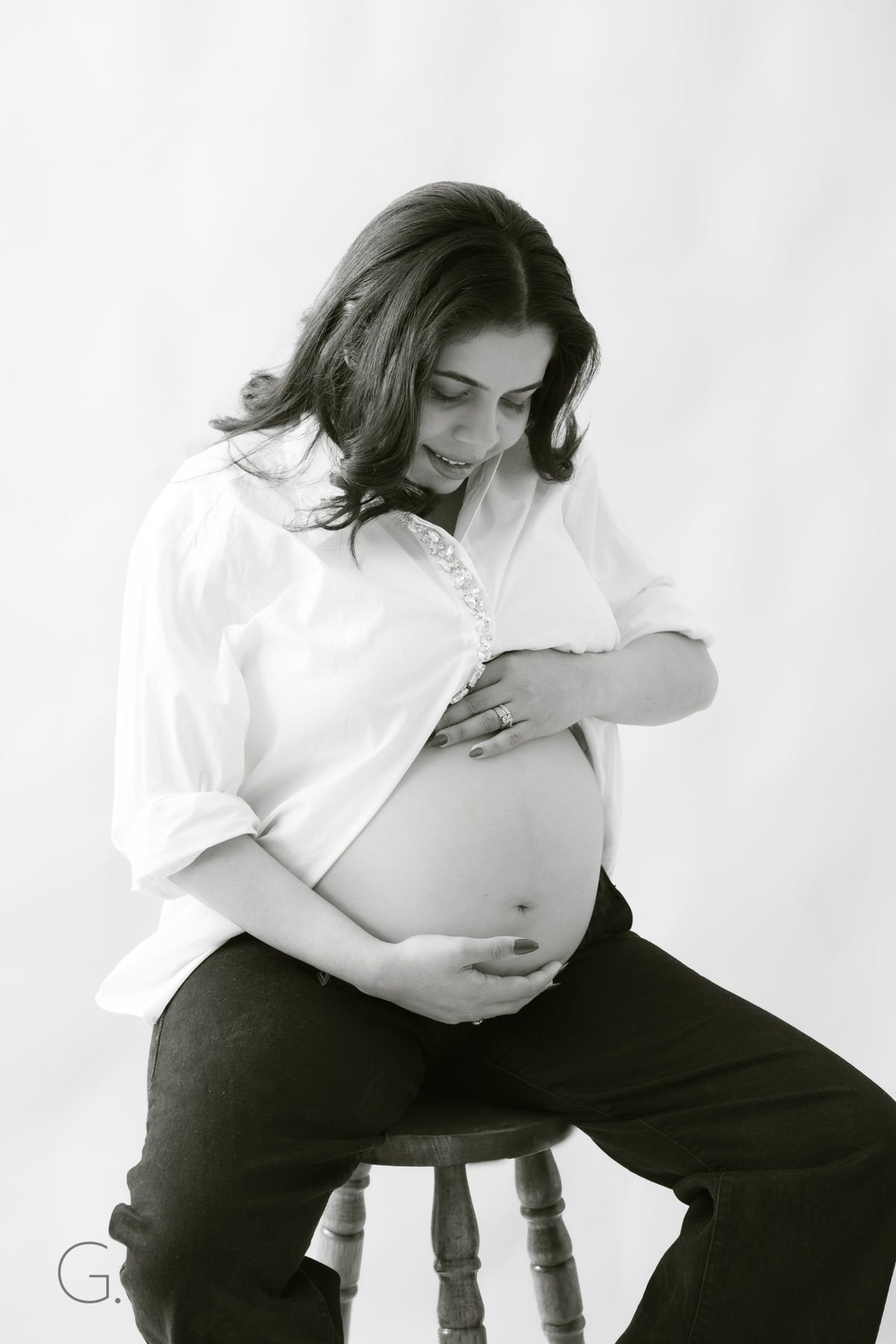 Black and white maternity portrait on a wooden stool, both hands on the bump
