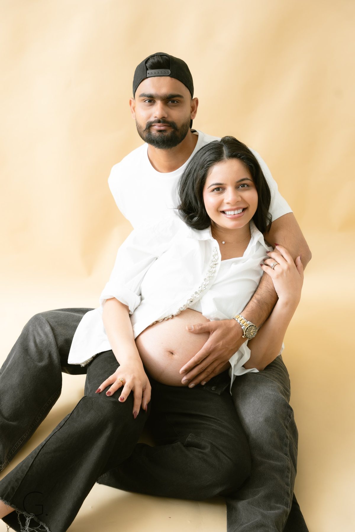 Couple seated on the studio floor, his hand on her bump, both smiling to camera — couples maternity shoot Melbourne