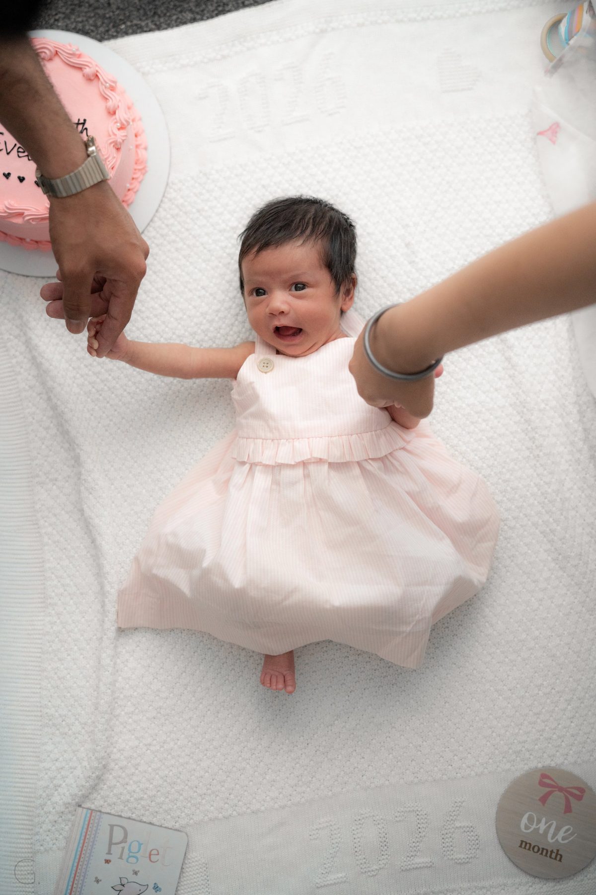 One-month-old in a pink dress on a knitted blanket, holding her parents' hands beside a 'one month' marker — at-home newborn photography